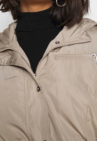 Béžová péřová bunda se zipem a patentkami, s kapucí a malou zipovou kapsou na hrudi, v kombinaci s černým rolákem.
