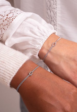 Silberdrahtarmbänder an zwei Handgelenken, mit einem einfachen Knotendesign. Die Armbänder sind mit strukturierten, hellen Ärmeln kombiniert.