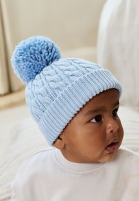 Lichtblauwe gebreide muts met grote pompon bovenop, voorzien van een kabelgebreid patroon en een geribbelde boord.