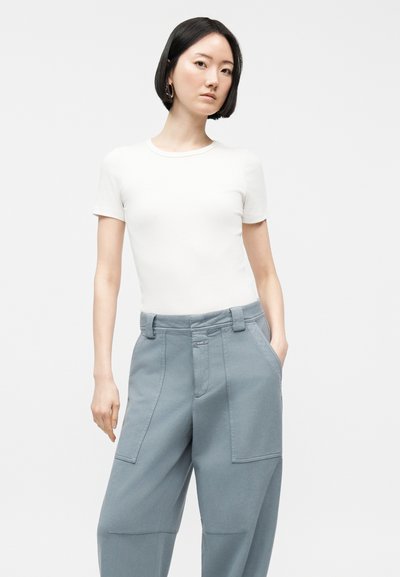 Γυναίκα με κοντά μαύρα μαλλιά, ντυμένη με εφαρμοστό λευκό πουκάμισο με κοντά μανίκια και φαρδιά ανοιχτό μπλε παντελόνια, όρθια με το ένα χέρι στην τσέπη.