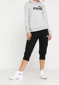 Persona vistiendo una sudadera gris de Puma, pantalones deportivos cortos negros de Puma y zapatillas deportivas blancas, de pie con una pierna ligeramente doblada.