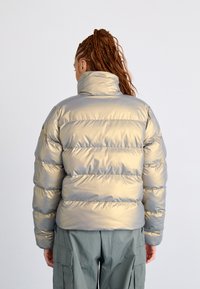 Giacca a vento metallica argento con design trapuntato orizzontale, colletto alto e trama liscia. Abbinata a pantaloni cargo verde oliva.