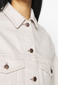 Giacca in denim beige con scollo a collo, cuciture visibili e bottoni in bronzo, con tasche sul petto e finitura morbida e strutturata.