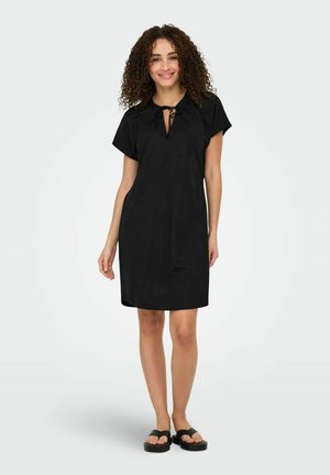 Robe noire à manches courtes avec un col à nouer, coupe droite et texture douce. Portée avec des sandales noires, elle présente un design épuré.