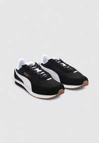 Para czarnych i białych sneakersów Puma z gumowymi podeszwami, białym paskiem z logo oraz czarnymi sznurówkami na jednolitym jasnym tle.