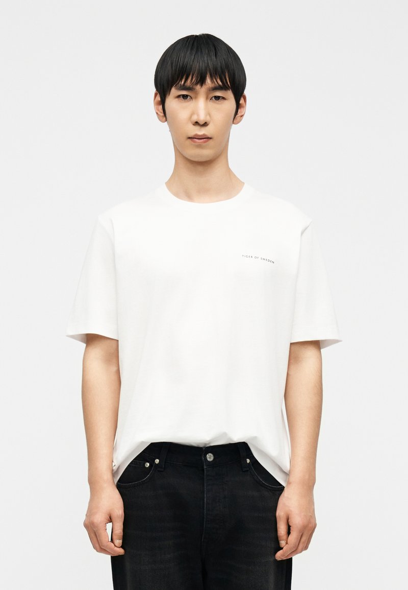 Νεαρός με ίσια μαύρα μαλλιά που φοράει απλό λευκ ό T-shirt με μικρό κείμενο και μαύρα τζιν, στέκεται μπροστά σε λευκό φόντο.