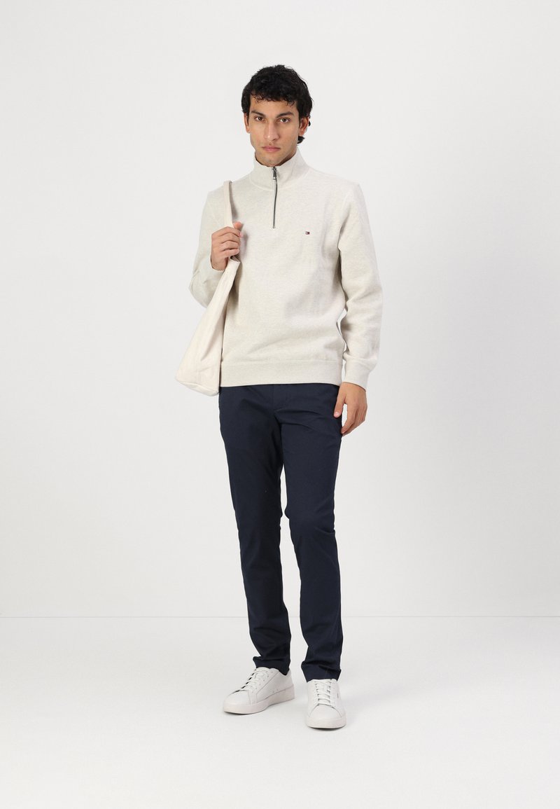 Lichtgrijze hoodie met ritssluiting en ribbels aan de mouwen, marineblauwe broek en witte sneakers. Model houdt een lichte tote bag vast en staat tegen een effen achtergrond.