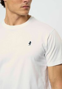 Balta medvilninė marškinė su mažai tamsiai mėlyna siuvinėta detale kairėje krūtinės pusėje. Apvalus iškirpimas ir trumpomis rankovėmis, lygus paviršius.