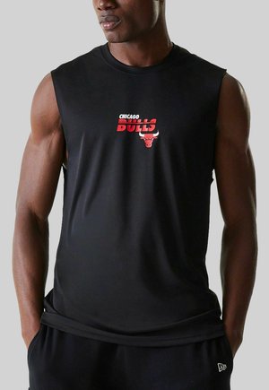 Mann in ärmellosem schwarzem Shirt mit rotem Chicago Bulls-Logo, der mit den Händen in den Taschen vor einem schlichten Hintergrund steht.