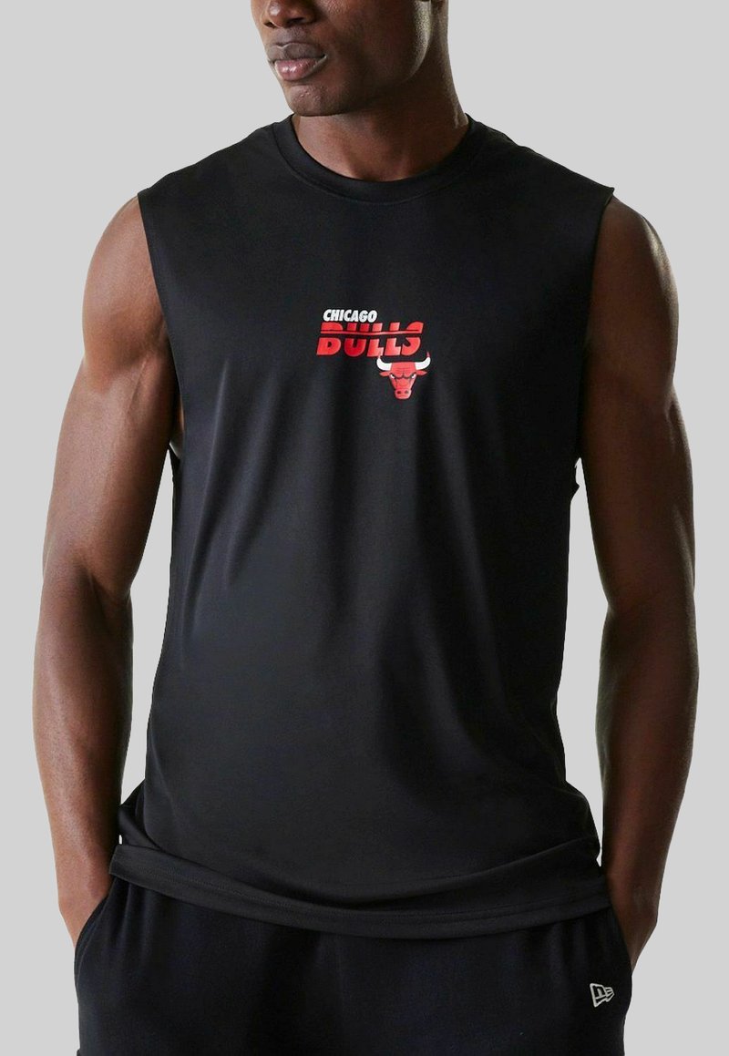 Mann in ärmellosem schwarzem Shirt mit rotem Chicago Bulls-Logo, der mit den Händen in den Taschen vor einem schlichten Hintergrund steht.
