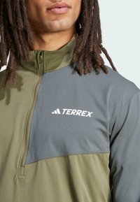 Giacca a pullover con zip al quarti in tessuto verde e grigio con una superficie liscia. Caratterizzata dal logo "TERREX" in bianco sul petto.