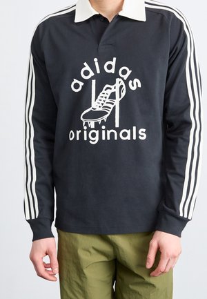 Schwarzes Polo mit langen Ärmeln, einem weißen Kragen und drei weißen Streifen an den Ärmeln, mit einem großen Grafikmotiv eines Schuhs und dem Text "adidas originals."