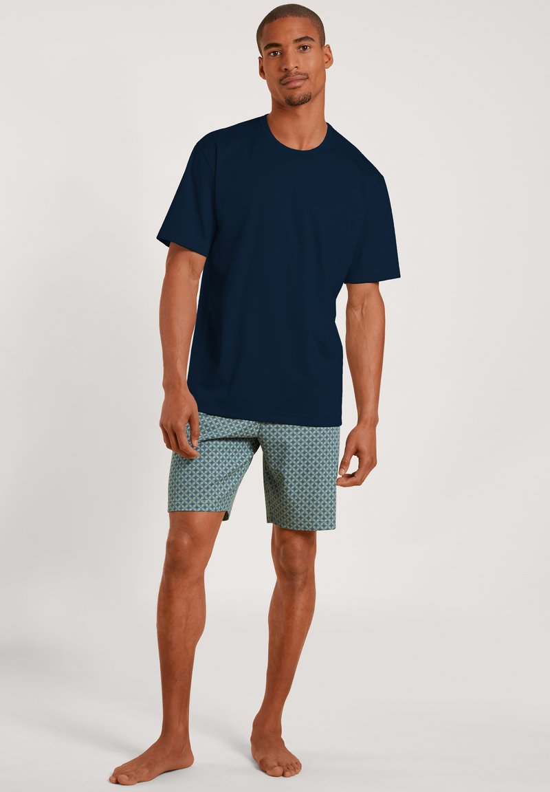 Marineblauw T-shirt met korte mouwen, gecombineerd met lichtgroene gestreepte shorts met een geometrisch patroon. Egaal achtergrond.
