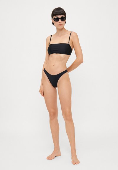 Melnais bikini komplekts, kas sastāv no bandeau tipa topa ar plānām siksnām un zemu jostasvietu biksītēm. Gluda auduma un minimālu dizaina elementu kombinācija.