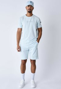 Lichtblauwe sportoutfit bestaande uit een t-shirt met korte mouwen en bijpassende shorts, gemaakt van een zachte stof met logovakken. Witte sneakers en sokken.