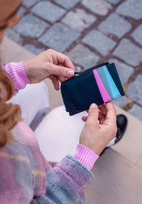 Compact black wallet with a zip closure, featuring vertical panels in pink, blue, and grey, held in a hand with a textured sweater.