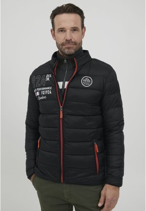 Chaqueta acolchada negra con cremallera naranja, diseño acolchado, varios parches y bolsillos laterales. Lleva puesta una camiseta de cuello alto gris.