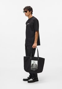 Črna platnena torba z usnjenimi ročaji, ki prikazuje natisnjeno podobo moškega v obleki. Torba ima veliko, strukturirano obliko.