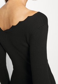 Haut en maille côtelée noire avec un design épaules dénudées festonné, manches longues et finition texturée.