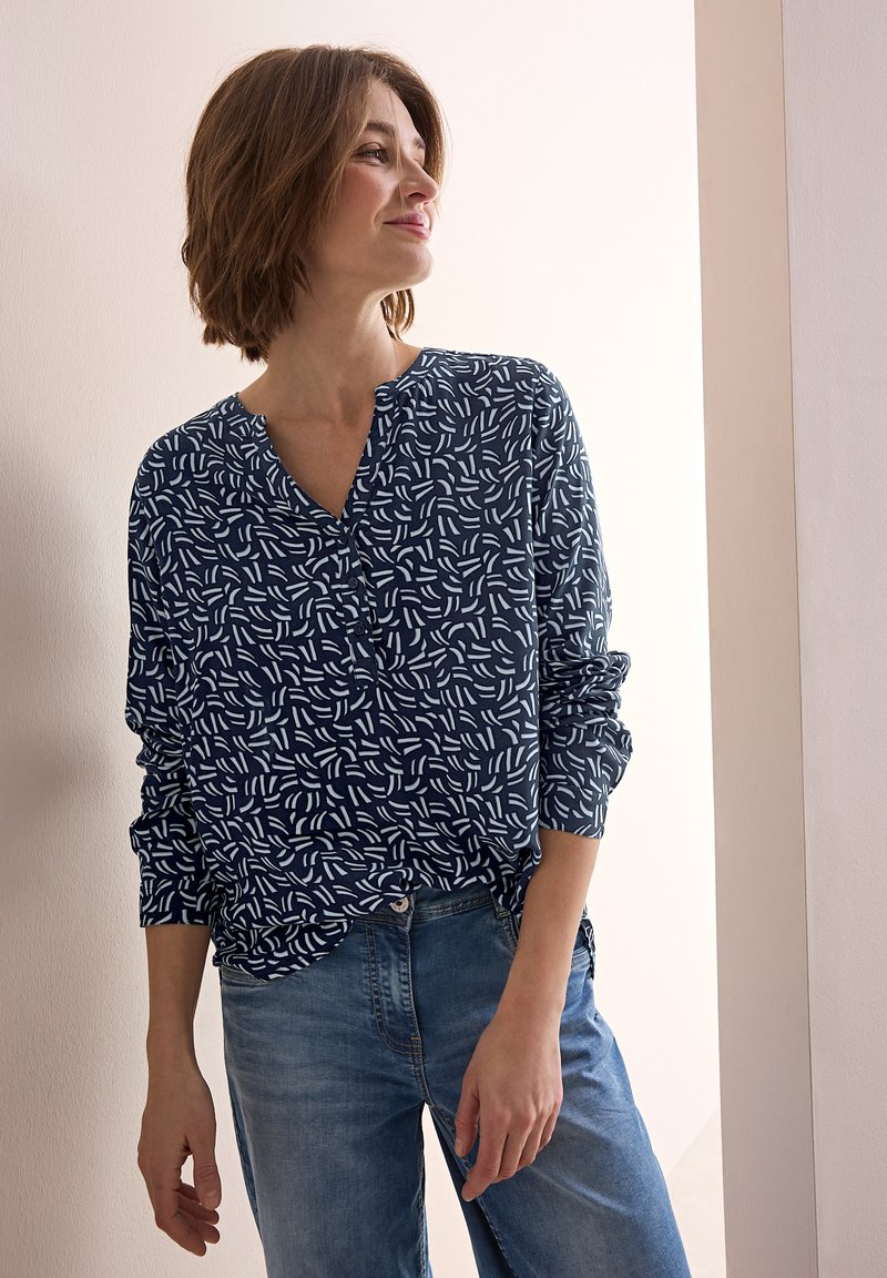 Marinblus med vitt abstrakt mönster, V-ringning, långa ärmar och mjukt tyg, kombinerad med ljusblå denimjeans.