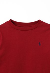 Röd bomulls-T-shirt med rund halsringning och en liten blå logotyp på vänster bröst. Mjuk textur, korta ärmar och enkel design.