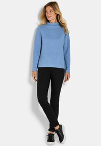 Maglietta azzurro chiaro a maniche lunghe con scollatura alta e tessuto testurizzato, abbinata a pantaloni neri aderenti e sneakers nere con suola bianca.