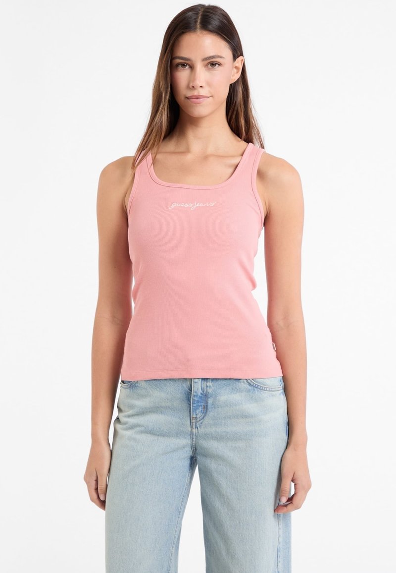 Roza rebrasta majica bez rukava sa kvadratnim izrezom, na kojoj se nalazi "guessjeans" logo u bijeloj boji, u kombinaciji s svijetloplavim širokim farmerkama.
