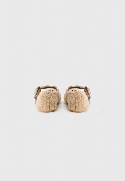Fuzzy beige backless slippers with a textured exterior, a rounded shape, and a decorative buckle strap in brown. Soft, cozy design.