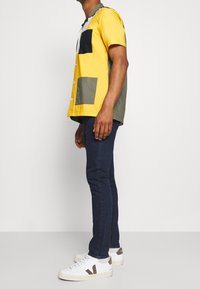 Camisa de manga corta con paneles en amarillo, verde y marino, con botones en la parte delantera y dos grandes bolsillos. Combinada con jeans oscuros y zapatillas blancas.