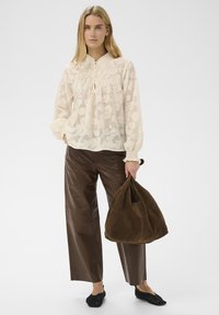 Femme aux longs cheveux blonds portant un chemisier crème transparent et un pantalon en cuir marron, tenant un grand sac en daim marron, debout sur un fond blanc.