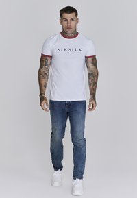 Witte crewneck t-shirt met rode afwerking, featuring zwart "SIKSILK" logo; gecombineerd met blauwe spijkerbroek en witte sneakers.