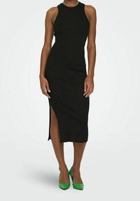 Robe midi noire à côtes avec un col haut et une fente sur le côté. Associée à des escarpins pointus verts vif. Texture lisse et design ajusté.