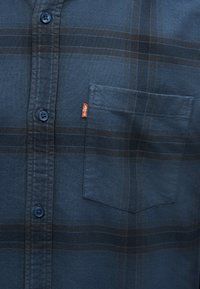 Σκούρο μπλε πουκάμισο με καρό σχέδιο, μπροστινή τσέπη και εμφανές ετικέτα Levi's στο πλάι της τσέπης, κλείσιμο με κουμπιά μπροστά με μπλε κουμπιά.