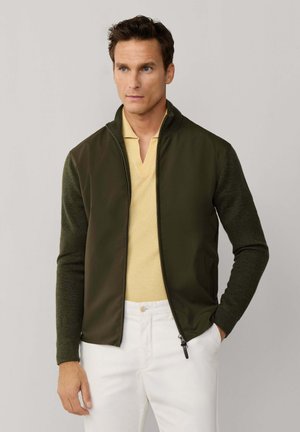 Hombre con chaqueta verde oliva con cremallera encima de una camisa tipo polo amarilla clara y pantalones blancos, de pie contra un fondo liso.