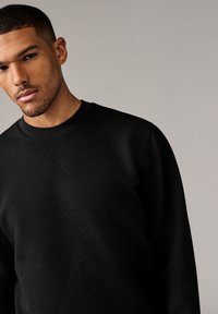 Felpa nera con trama a rilievo, con scollatura rotonda, caratterizzata da un motivo a diamante in rilievo e polsini a coste, che mette in evidenza un design aderente.