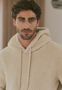 Beige fleece-huppari, jossa on rakenteellinen pinta, narut ja suuri huppu. Kangas näyttää pehmeältä ja lämpimältä, sopii rentoon käyttöön.