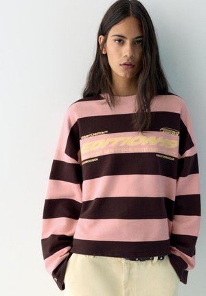 Jonge vrouw met lang donker haar, gekleed in een roze en donkerbruine sweater met brede strepen en gele tekst, staande tegen een witte muur.