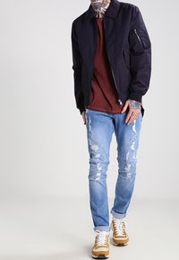 Marynarka lotnicza w kolorze granatowym, bordowa koszulka, jasnoniebieskie jeansy z przetarciami oraz beżowe sneakery z białymi sznurówkami. Widoczne tatuaże na ramionach.