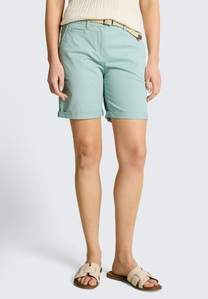 Kobieta w jasnoniebieskich szortach z podwiniętym brzegiem, beżowym plecionym paskiem, kremowym dzianinowym topem i beżowymi sandałami typu slide na jednolitym tle.