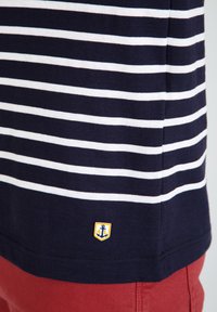 Marine og hvidstribet T-shirt med et lille gult emblem med et anker ved kanten, parret med røde shorts.