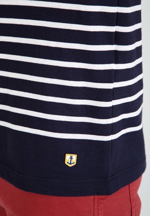T-shirt rayé bleu marine et blanc avec un petit emblème jaune représentant une ancre à l'ourlet, assorti de shorts rouges.