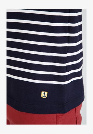T-shirt rayé bleu marine et blanc avec un petit emblème jaune représentant une ancre à l'ourlet, assorti de shorts rouges.