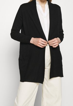 Personne attachant un long cardigan noir par-dessus une chemise blanche boutonnée et un pantalon large crème, visible du cou aux cuisses.