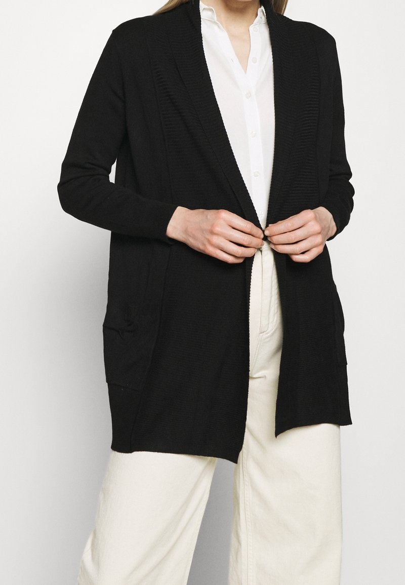 Personne attachant un long cardigan noir par-dessus une chemise blanche boutonnée et un pantalon large crème, visible du cou aux cuisses.