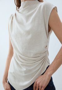 Haut sans manches beige clair avec un col montant et une texture drapée. Présente des détails froncés sur le côté et un tissu tissé lisse.
