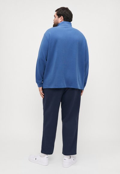 Blue, long-sleeve fleece top with a high collar; paired with dark navy pants and white sneakers. Textured fabric, relaxed fit, simple design.