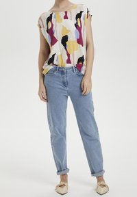 Blusa clara con patrones abstractos en amarillo, rosa y negro. Jeans de cintura alta azules, remangados en los puños, combinados con zapatos planos color nude.