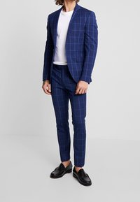 Marinblå rutiga kostym med skräddarsydd passform, som har en kavaj med en knapp och slim-fit byxor. Kombineras med en vit t-shirt och svarta loafers.