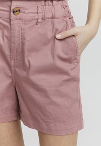 Lys rosa bomullsshorts med en avslappet passform, med sidelommer og knappelukking, synlig rynket tekstur.