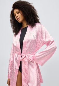 Ροζ σατέν ρόμπα με σχέδιο καρδιές, κλείσιμο με ζώνη και φαρδιά μανίκια. Διακριτική γυαλάδα με διακοσμητική καρδιά στο στήθος.